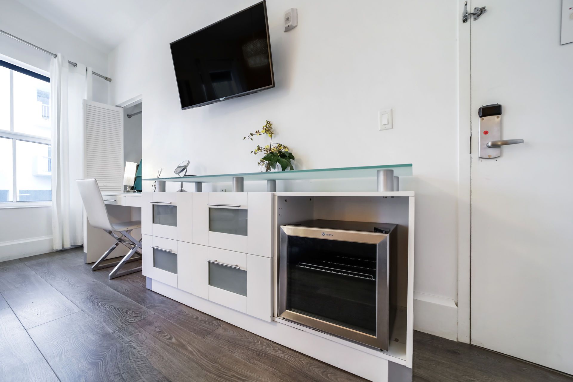 White room with desk, mini-fridge, and wall-mounted TV. Dark wood floor and white door.