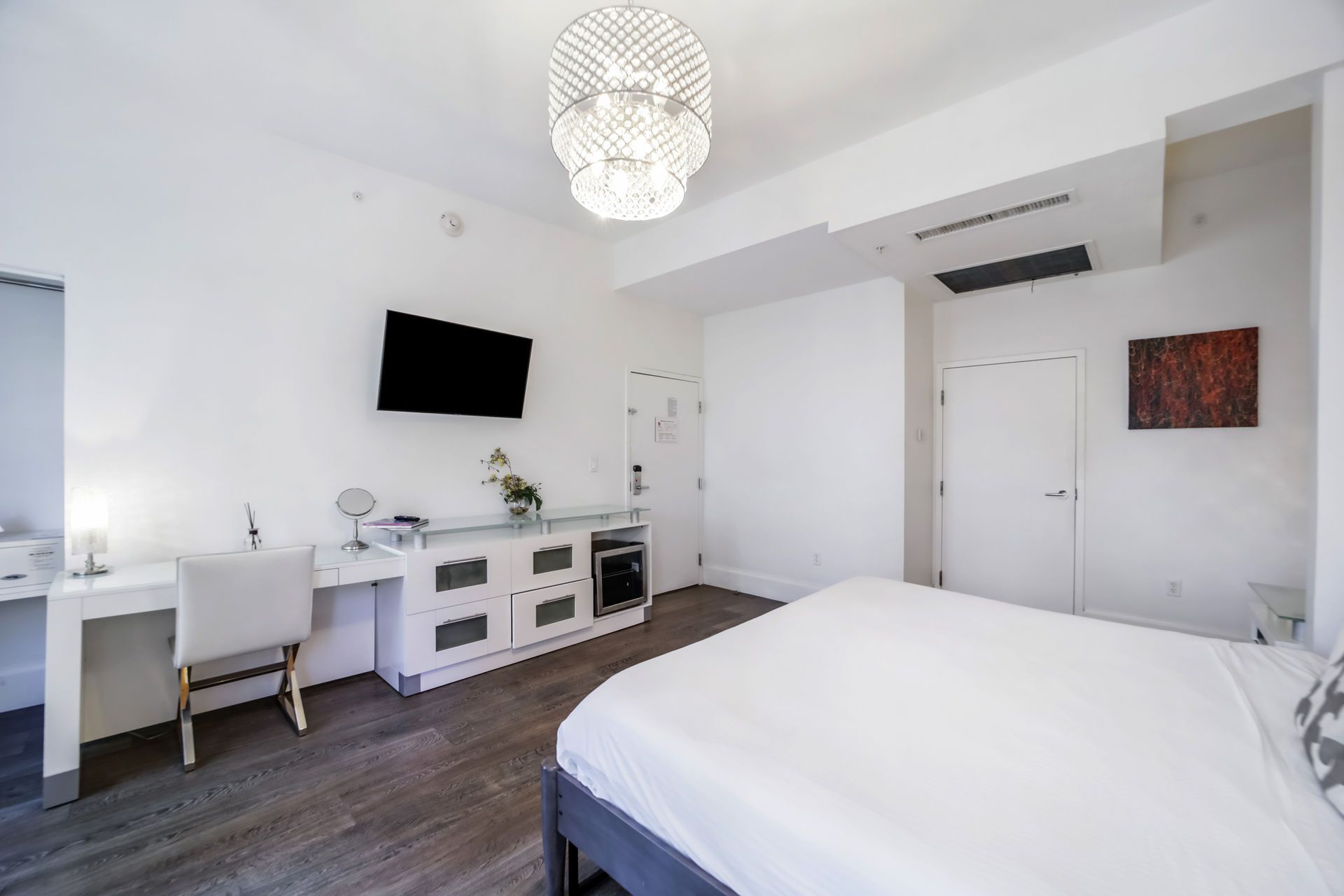 Bright hotel room with a white bed, desk, and built-in dresser. A TV is mounted on the wall.
