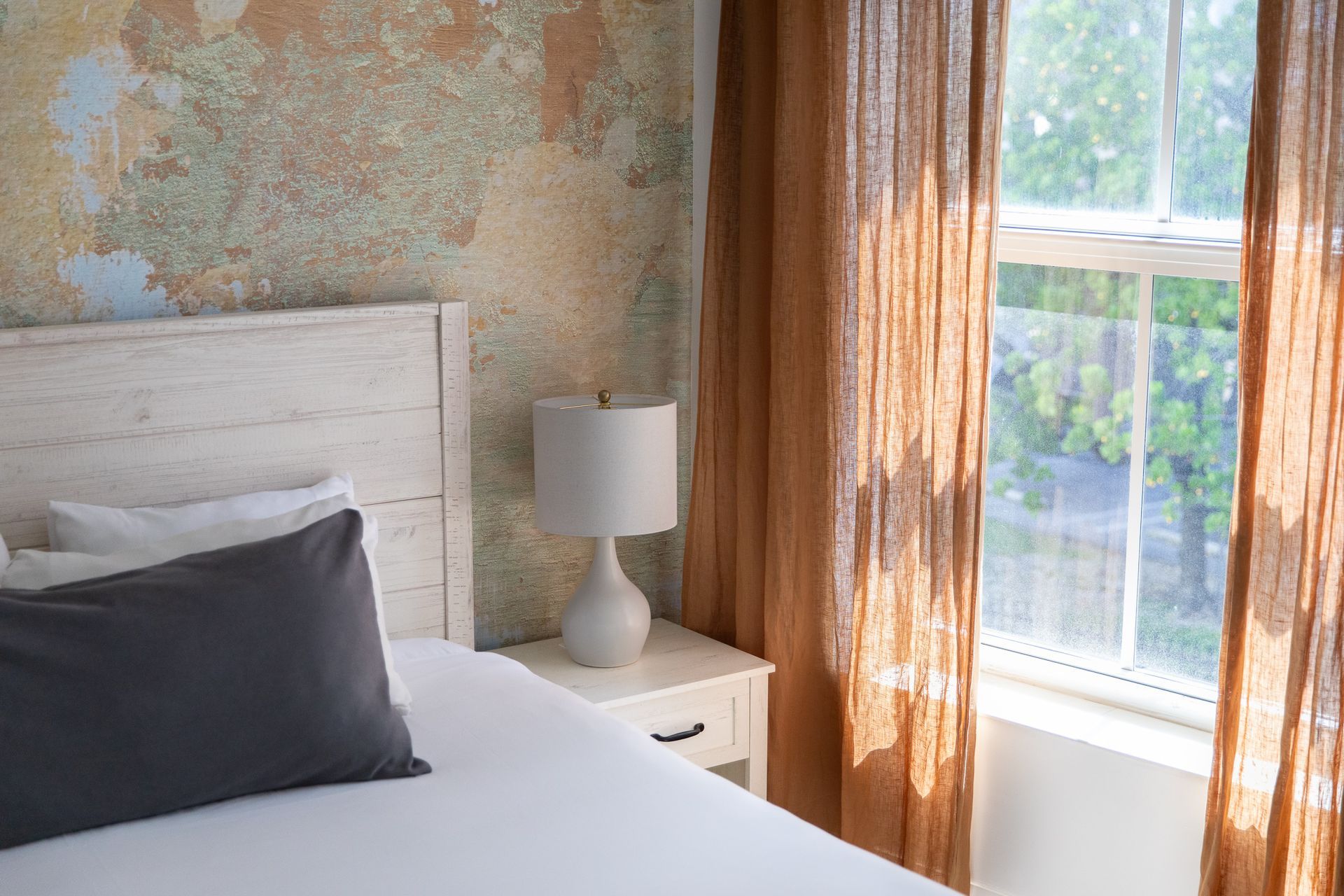 Bedroom with bed, nightstand lamp, and sunlit window with sheer curtains