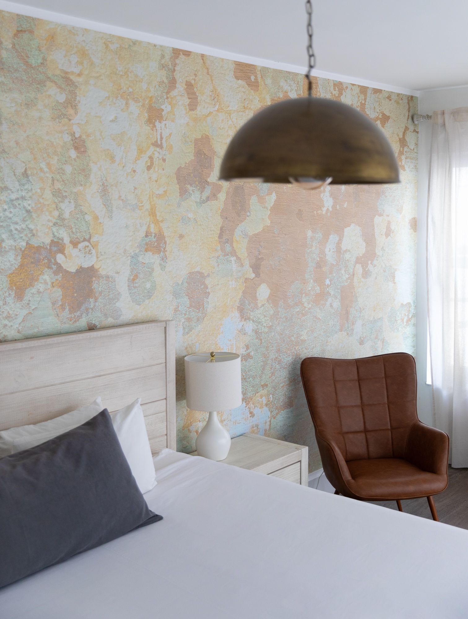 Modern bedroom with textured pastel wall, white bed, brown chair, bedside lamp, and pendant light