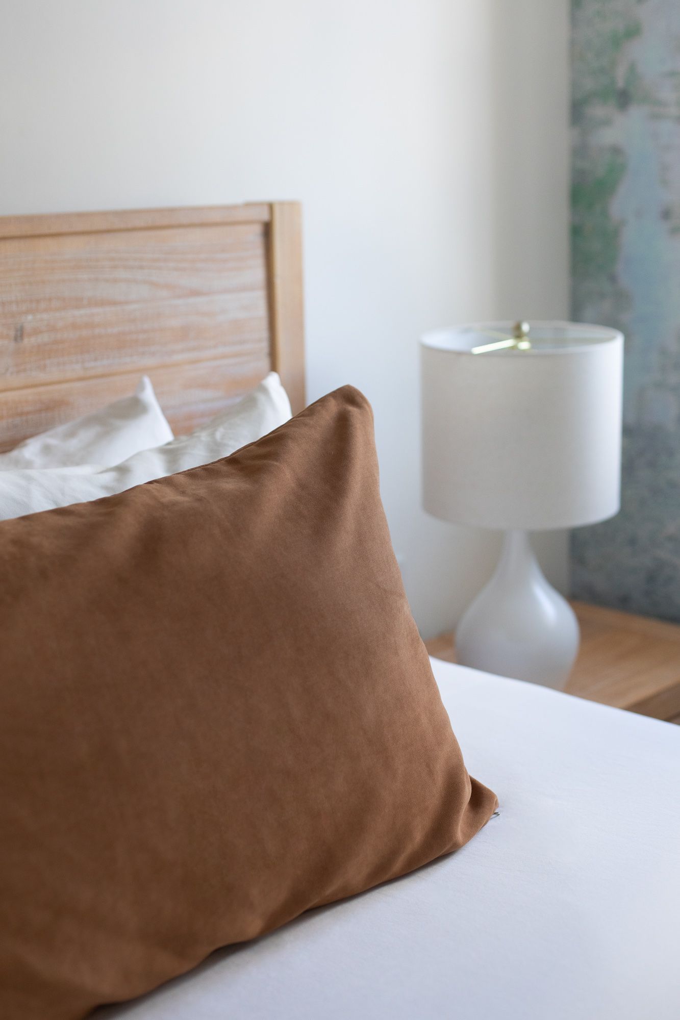 Brown pillow on a bed beside a white lamp on a wooden nightstand in a bright bedroom