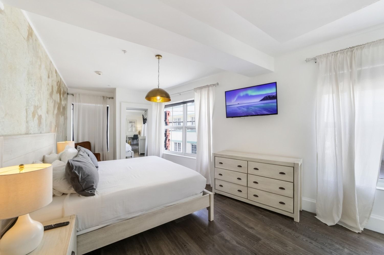 Bright white bedroom with a made bed, dresser, wall TV, and sheer curtains with city view.