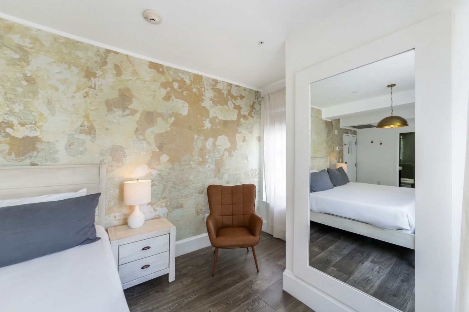 Bright bedroom with weathered floral wall, bed, bedside table, armchair, and large mirror wardrobe.