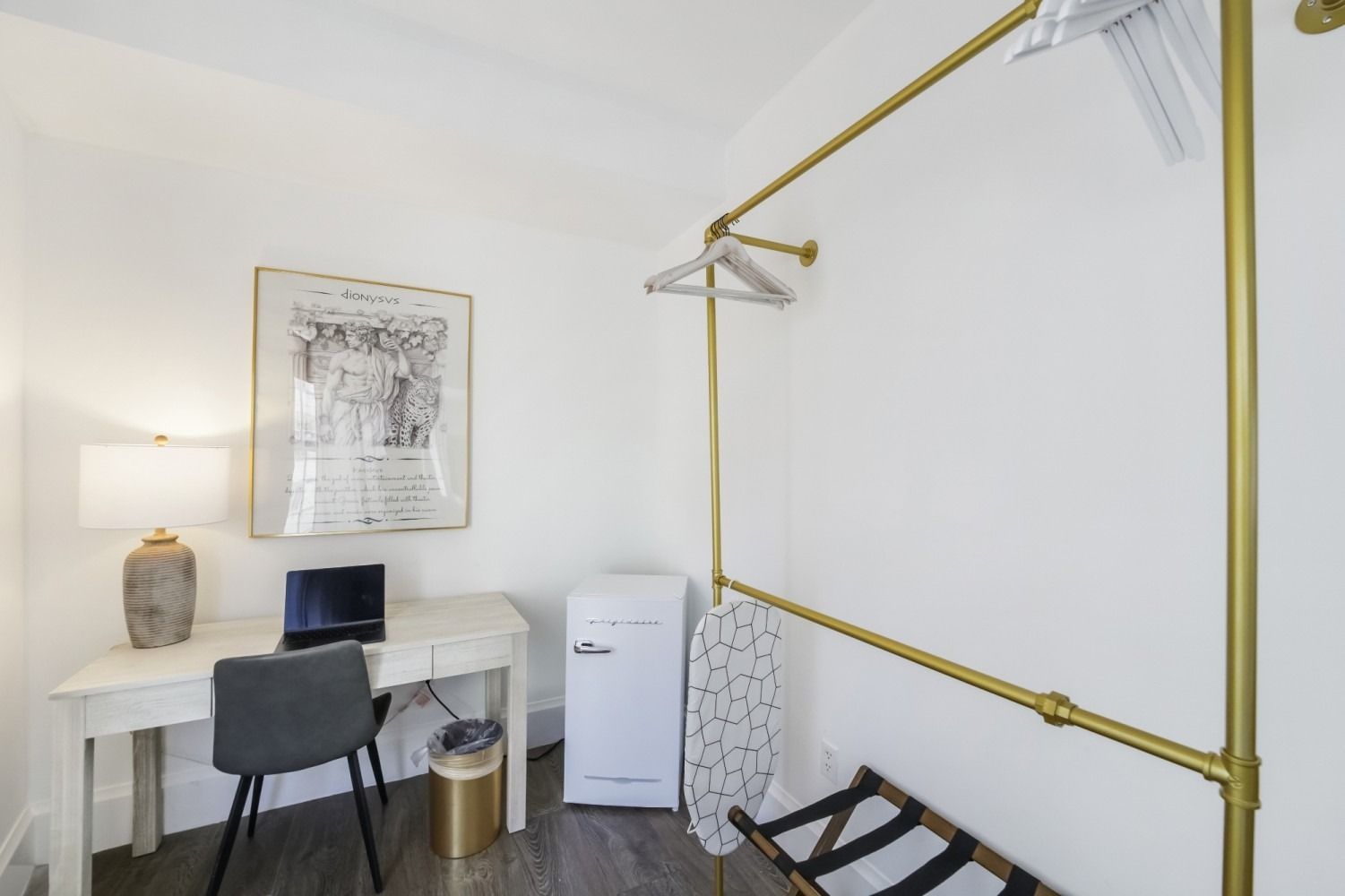 Minimal home office with desk, laptop, chair, lamp, mini fridge, and gold wall-mounted garment rack