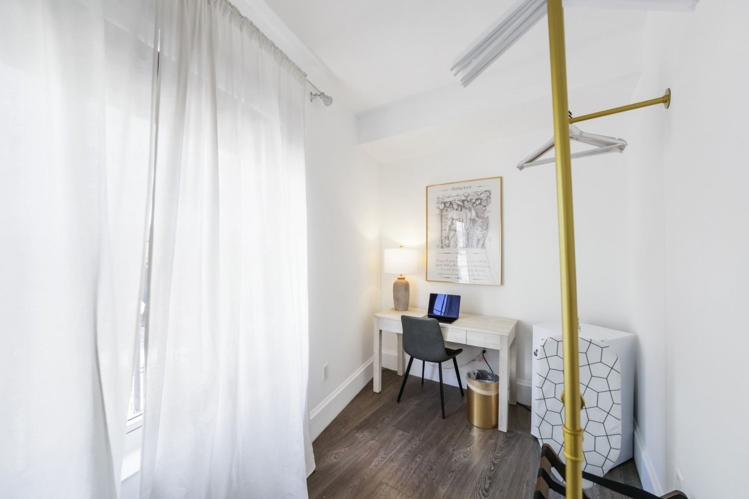 Bright white bedroom nook with desk, chair, lamp, wall art, and a gold clothing rack by a large curtain