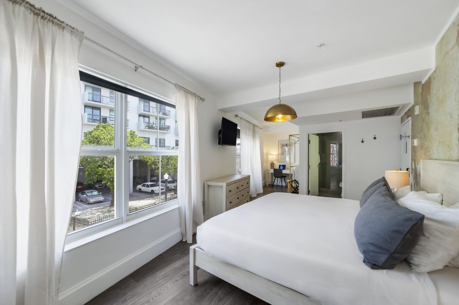 Bright bedroom with large window, white bedding, gray pillows, and a pendant light