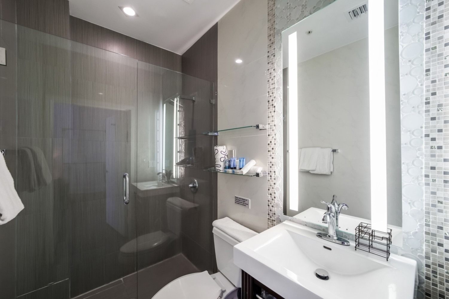 Modern bathroom with glass shower, floating sink, illuminated mirror, and mosaic tile accents