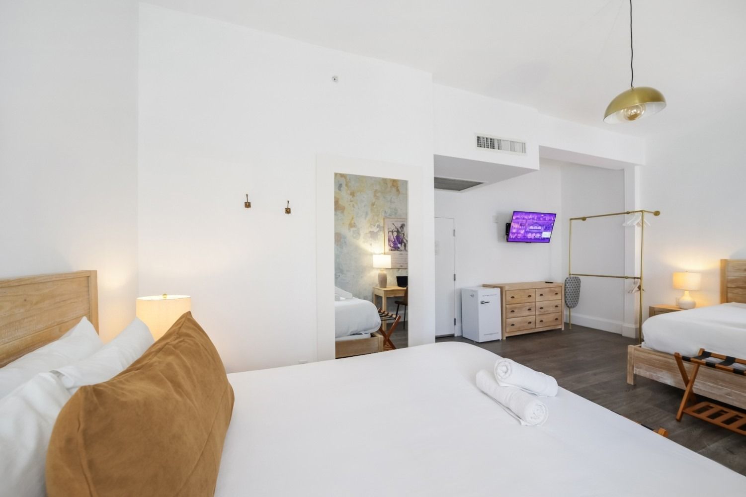 Bright white hotel room with two beds, a wall-mounted TV, and an open bathroom doorway