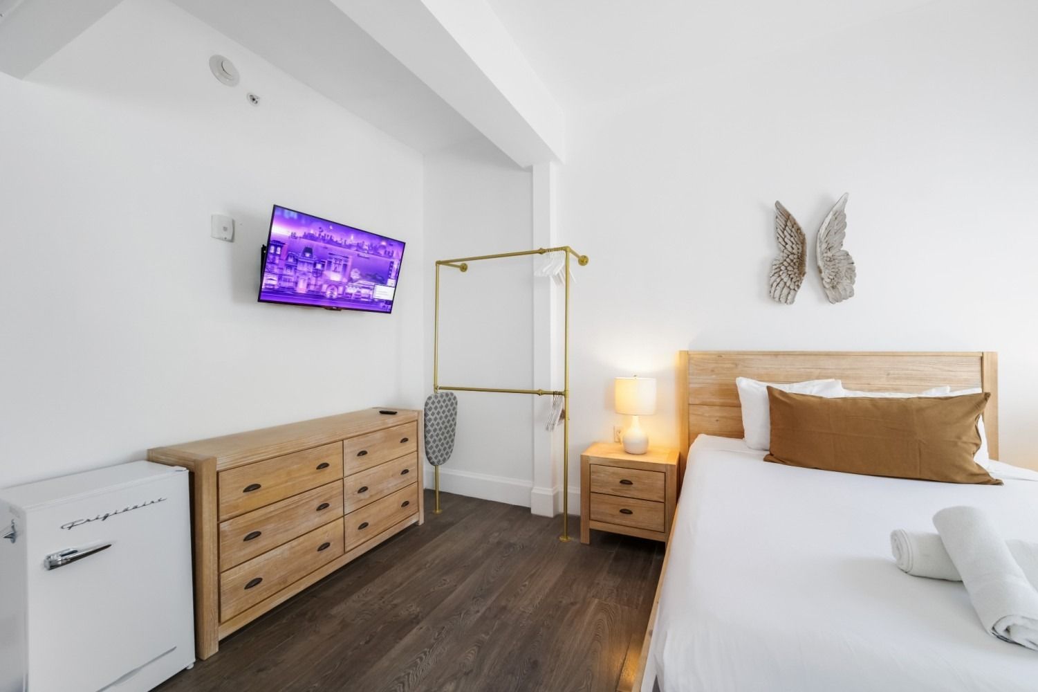 Bright minimalist bedroom with white walls, wood furniture, wall-mounted TV, and a bed with gold accents