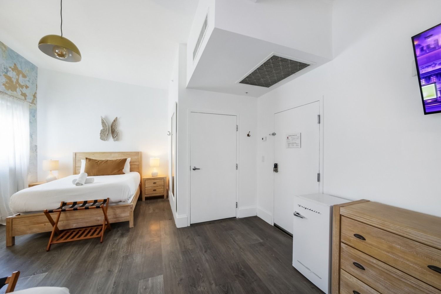 Bright modern bedroom with a bed, white walls, wood floors, dresser, and wall-mounted TV