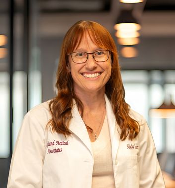 Smiling woman in a white lab coat and glasses, standing in a modern office or clinic setting.
