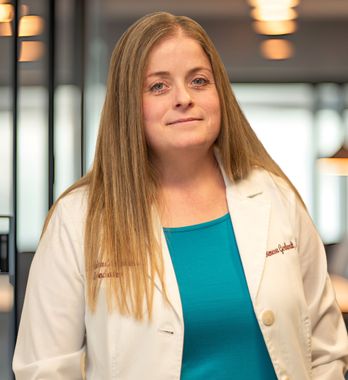 Woman in a white coat over a teal top, standing in a blurred indoor hallway.