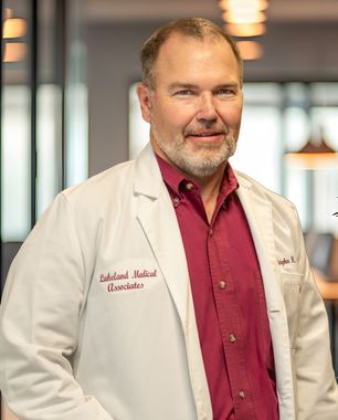 Doctor in a white coat and red shirt standing indoors, smiling at the camera.