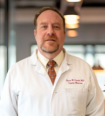 Portrait of a doctor in a white lab coat standing indoors, with a blurred hallway background