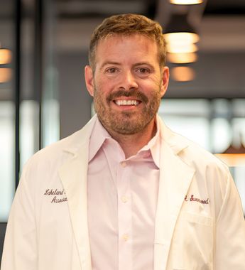 Smiling doctor in a white coat and pink shirt standing indoors.