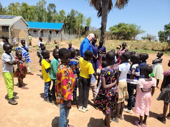 Children gather around a man in blue near a building with solar panels, under a palm tree.