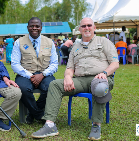 Two men seated outside smiling, wearing vests with a logo. One man is holding a hat and the other has a cane.