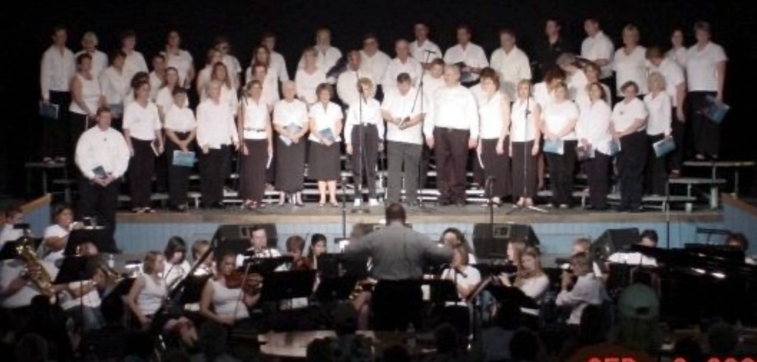Choir and orchestra performing on stage; conductor faces performers, who wear white shirts and dark bottoms.
