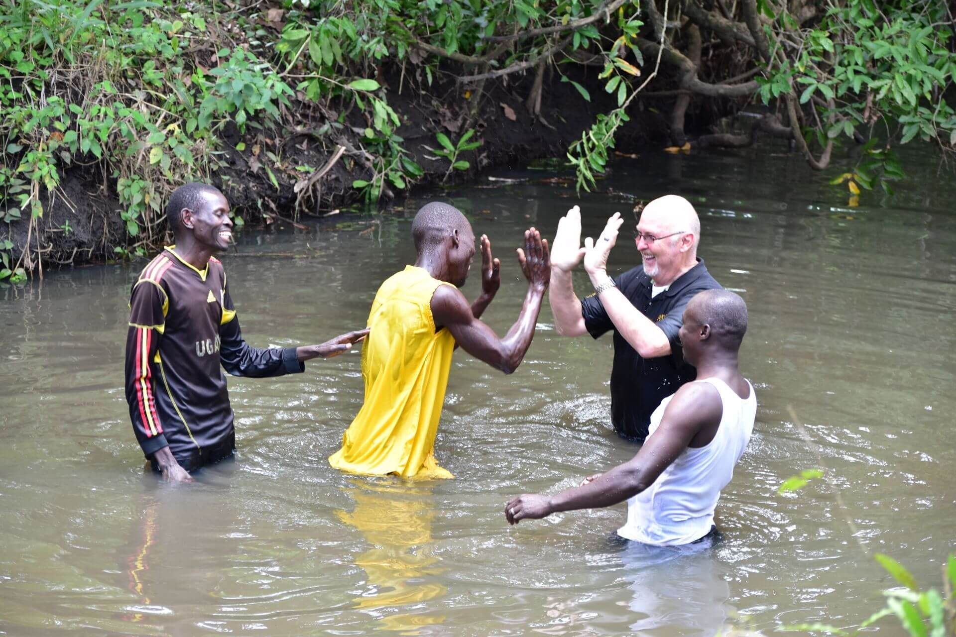 People in river giving high five, one wearing black shirt, others wet in yellow and white, surrounded by green foliage.