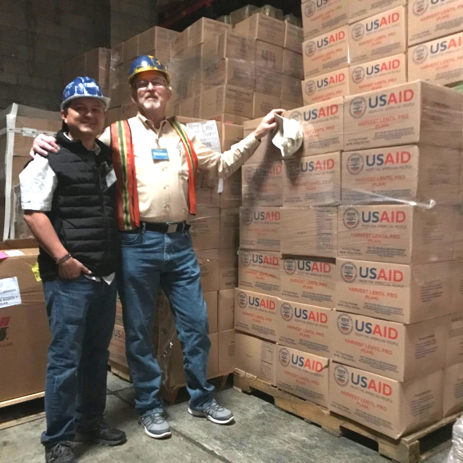 Two men posing with boxes of USAID food aid in a warehouse.