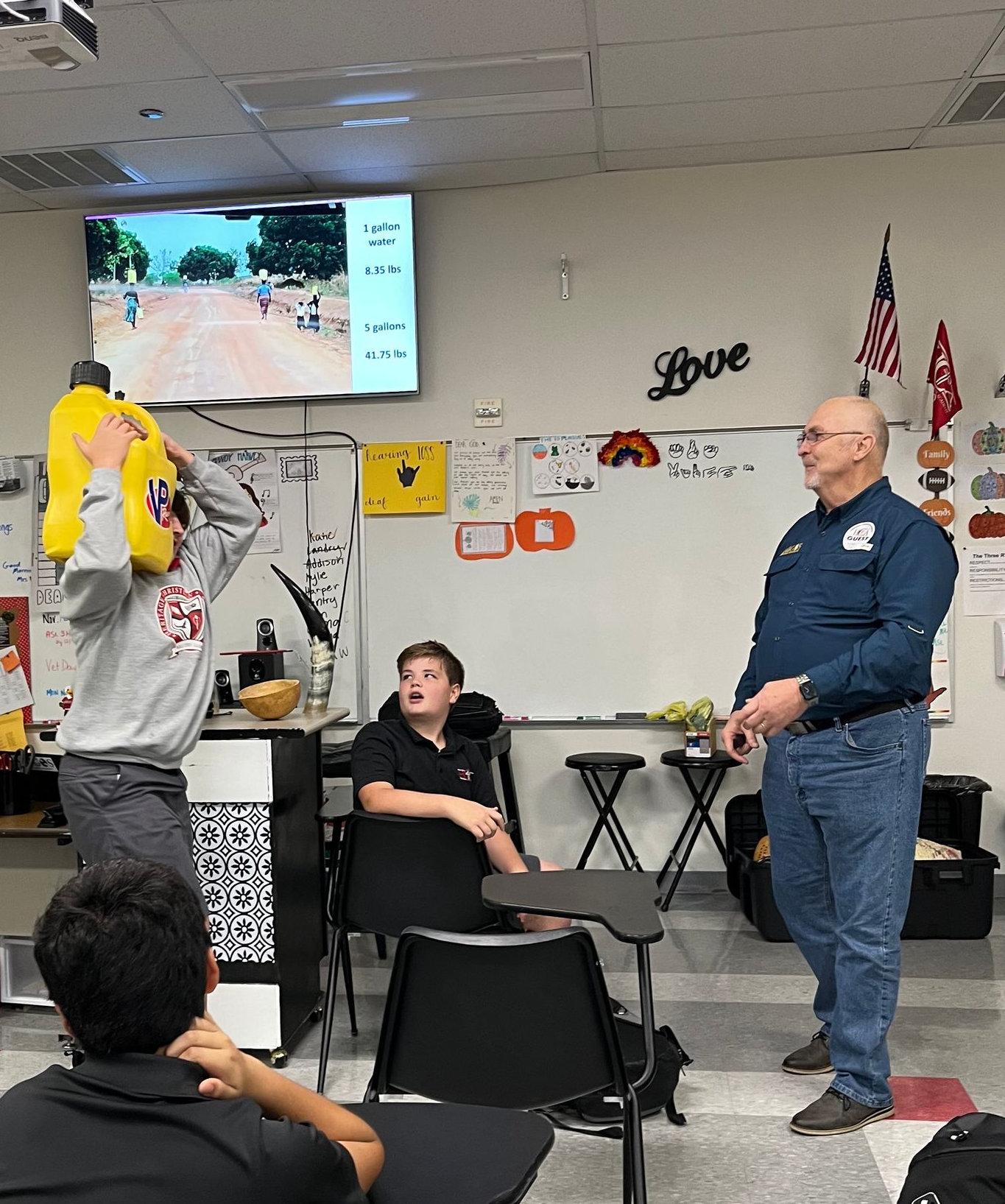 Students in classroom; one holds yellow object, another sits, a man stands. Whiteboard, screen visible.