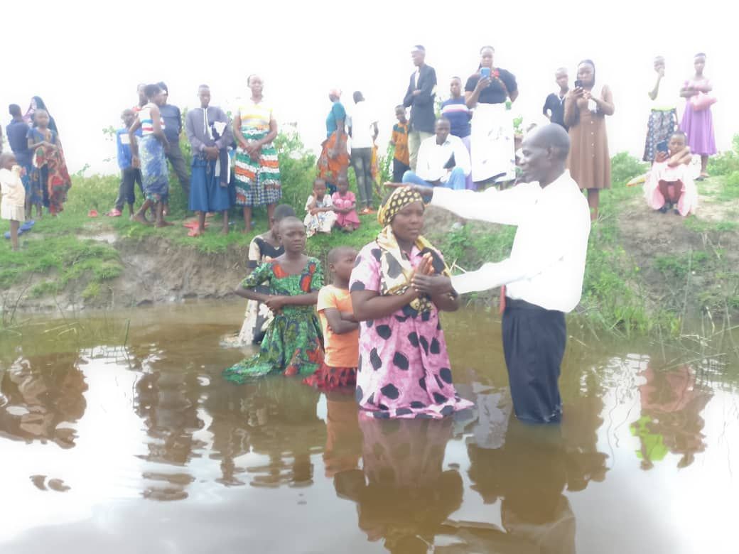 People participating in a baptism ceremony in shallow water; group of people watching.