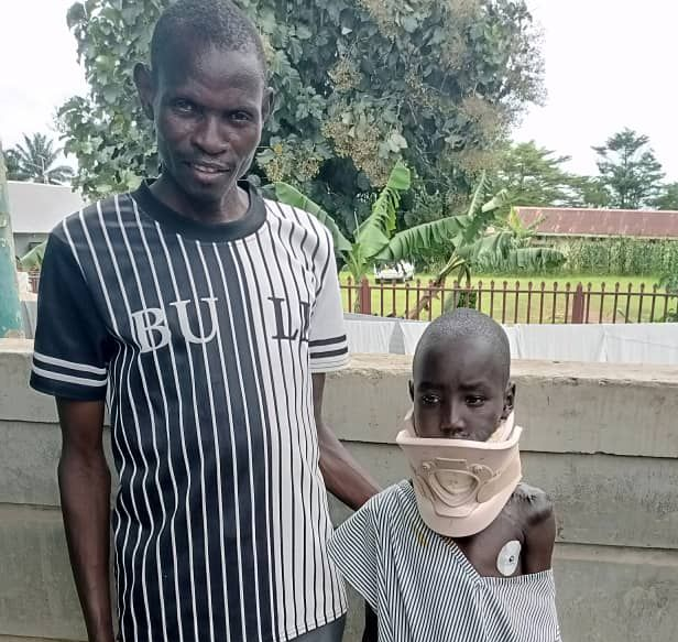 Man stands with a child wearing a neck brace and striped outfit on a patio.