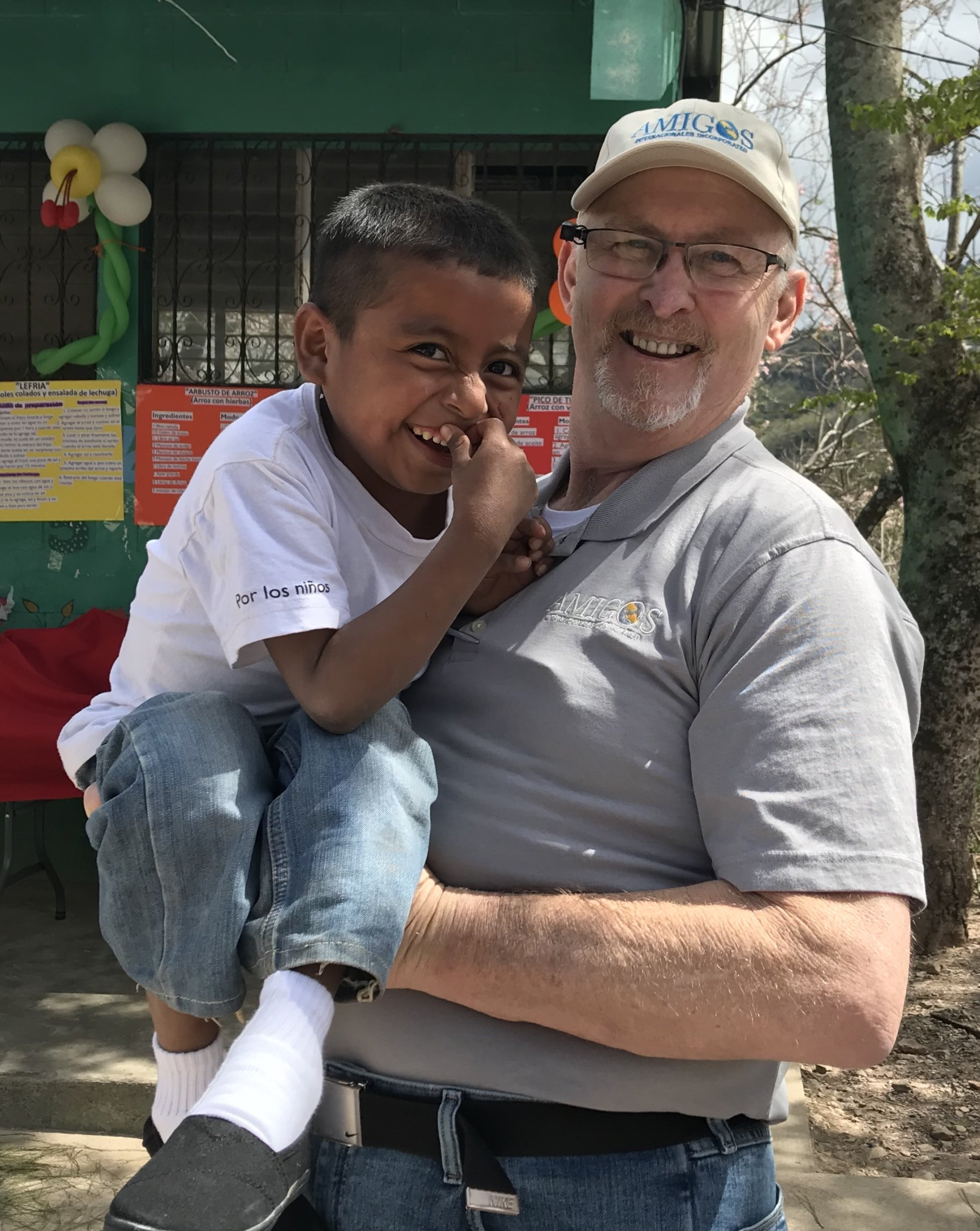 Man smiling, holding a child. Both appear happy. Outdoors, with a building in the background.