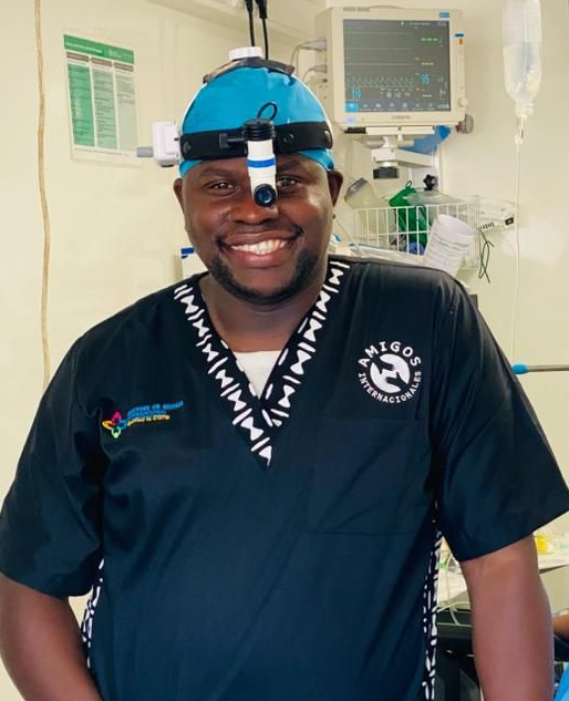 Surgeon in scrubs, headlight, and blue cap smiles in operating room setting.