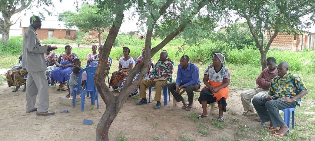 A group of people sitting under a tree listening to a man speaking. They are outdoors.