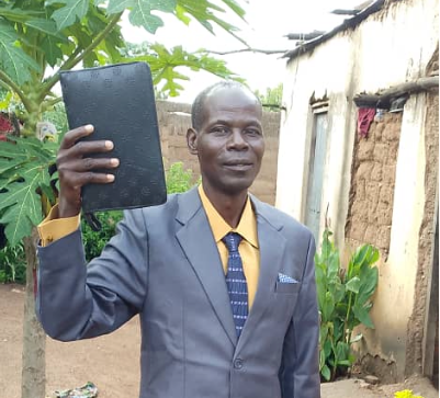 Man in suit holds up a dark book, smiling, outside a building with foliage.
