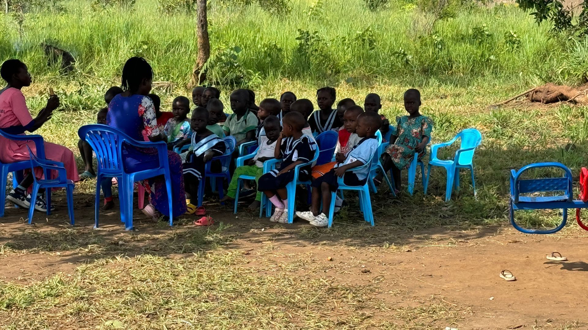 Children seated in chairs, listening to a woman in a park.