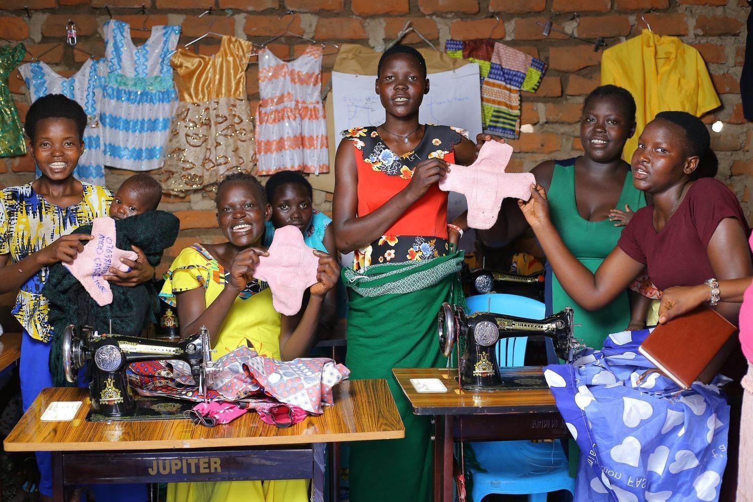 Women sewing reusable menstrual pads; various colorful outfits hang on the wall.