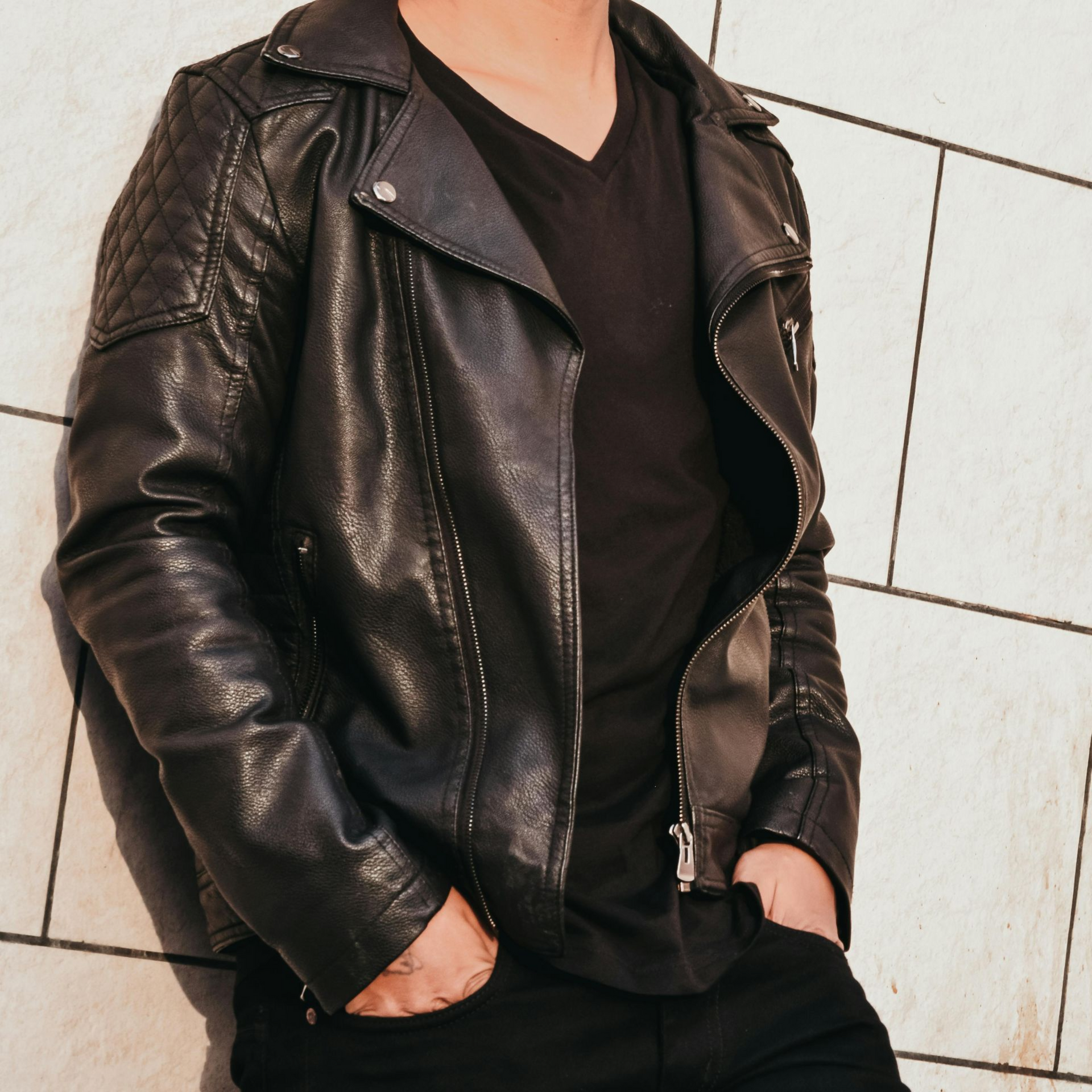 Person wearing a black leather jacket and black shirt, hands in pockets, leaning against a white tiled wall.