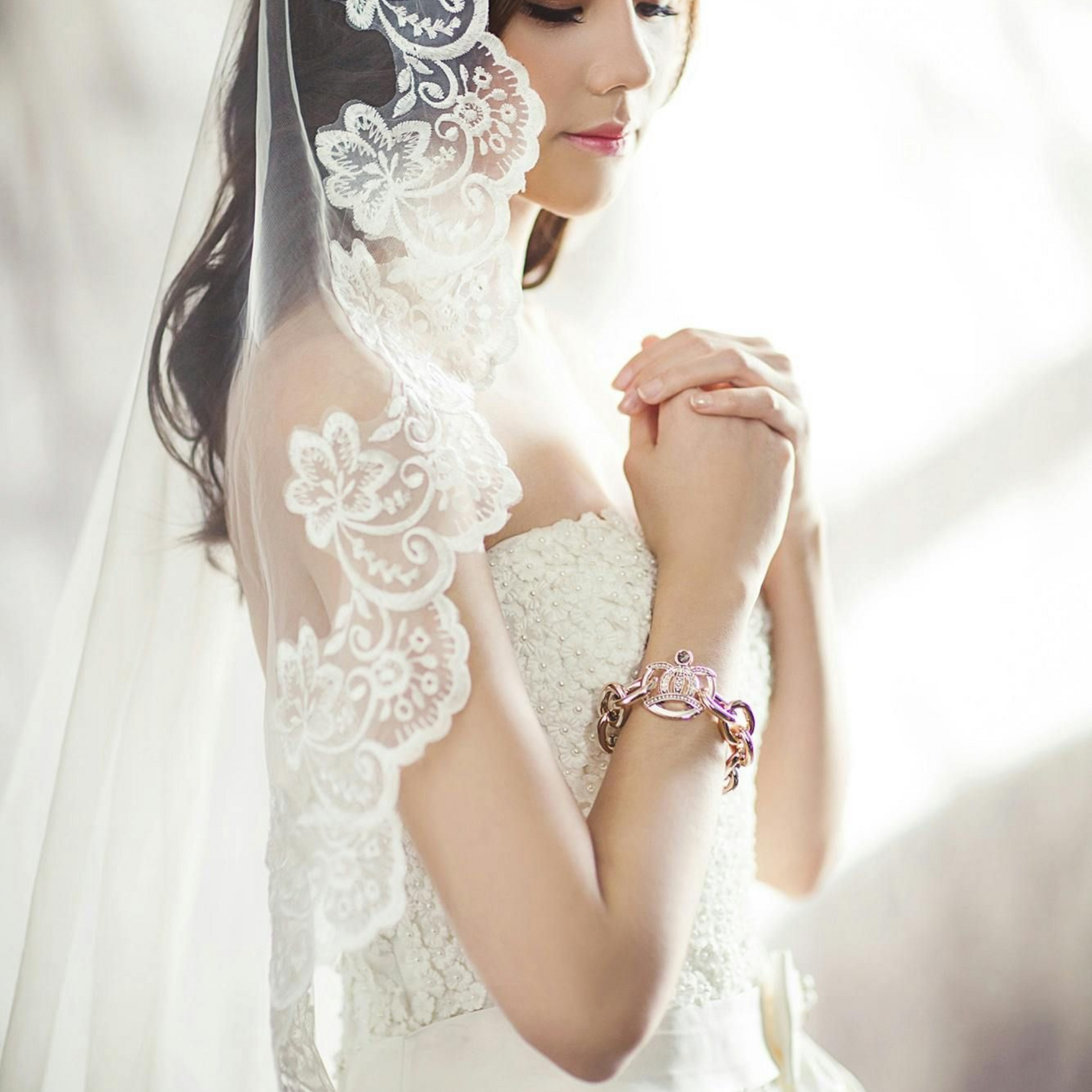 Bride in white lace veil and dress, hands clasped, sunlight.