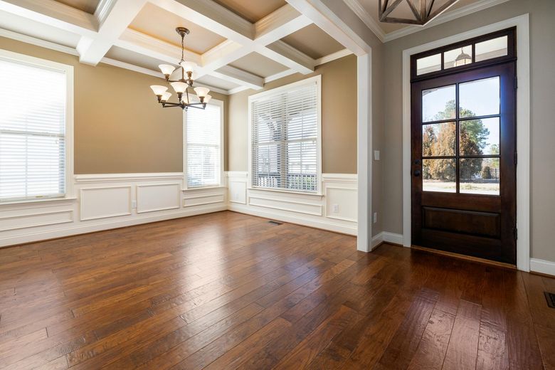 An empty dining room with coffered ceilings, light tan walls, white wainscoting, a chandelier, and a dark wood front door.