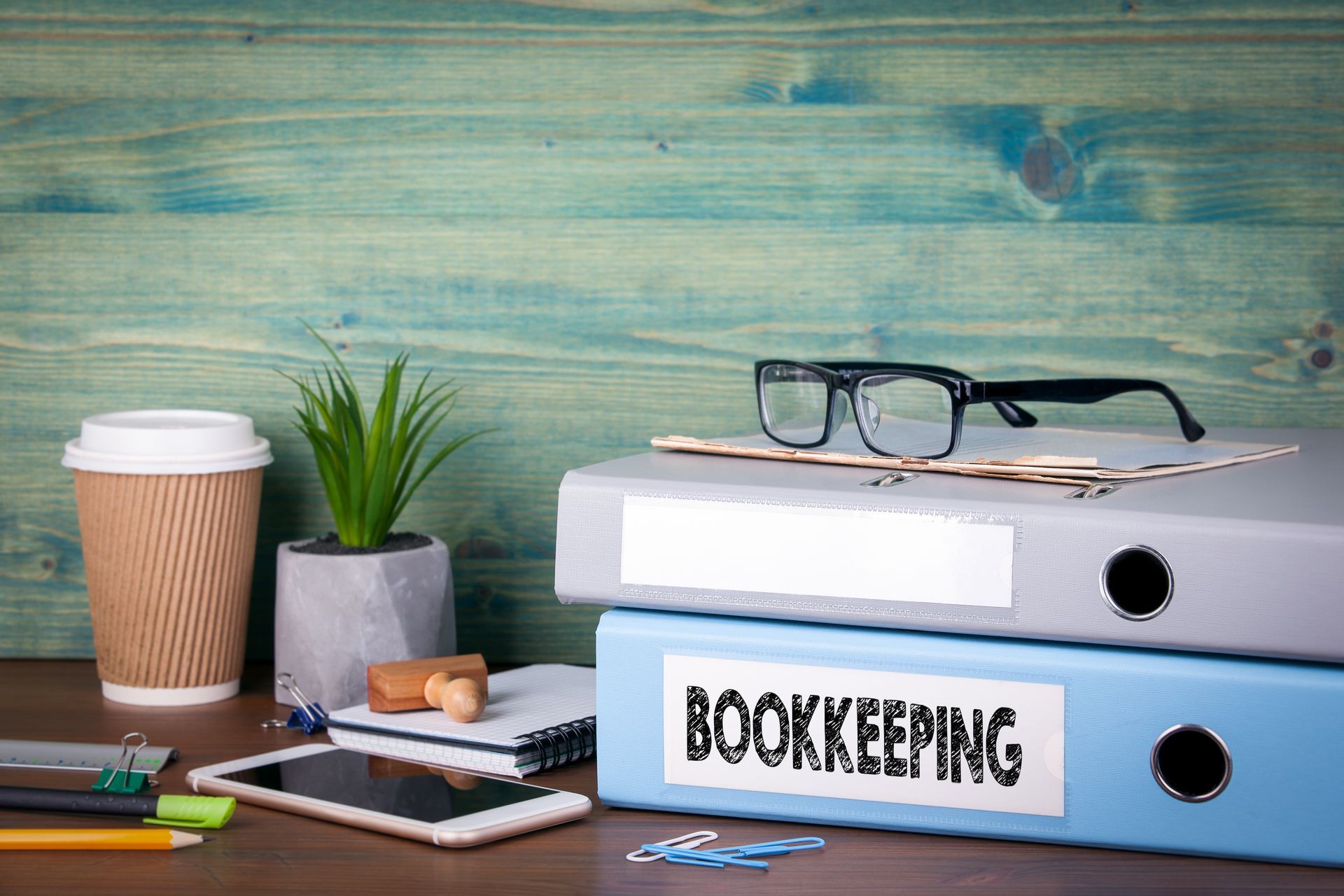 A neatly organised desk with a blue binder labelled 'Bookkeeping', a notebook, glasses, stationery, a smartphone, a coffee cup, and a small plant, symbolising professional and organised bookkeeping services.