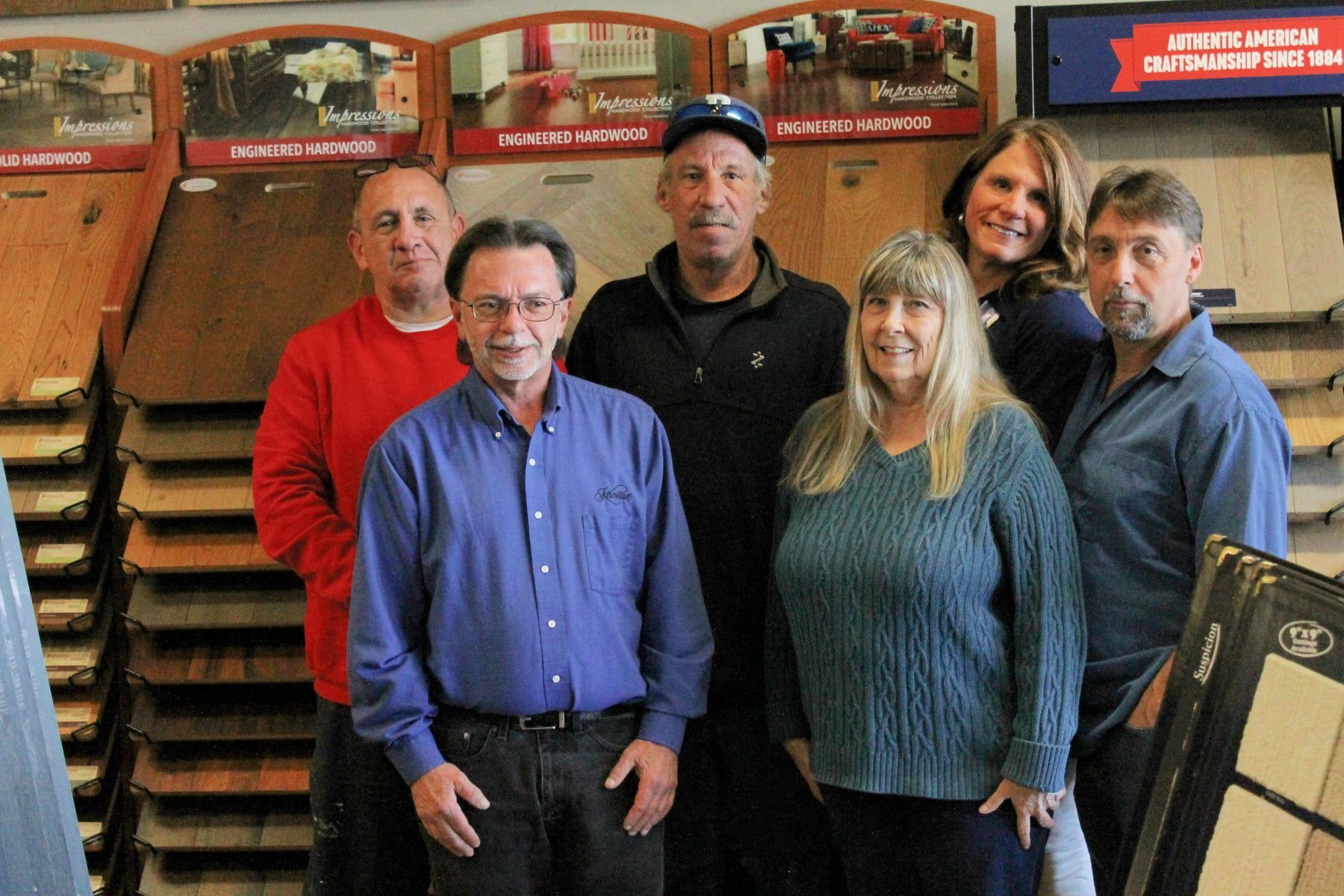 Group of six people smiling in a flooring store, standing in front of wood samples.