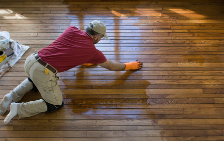 Man kneeling on hardwood floor, applying finish with orange gloves, wearing red shirt and khaki pants.