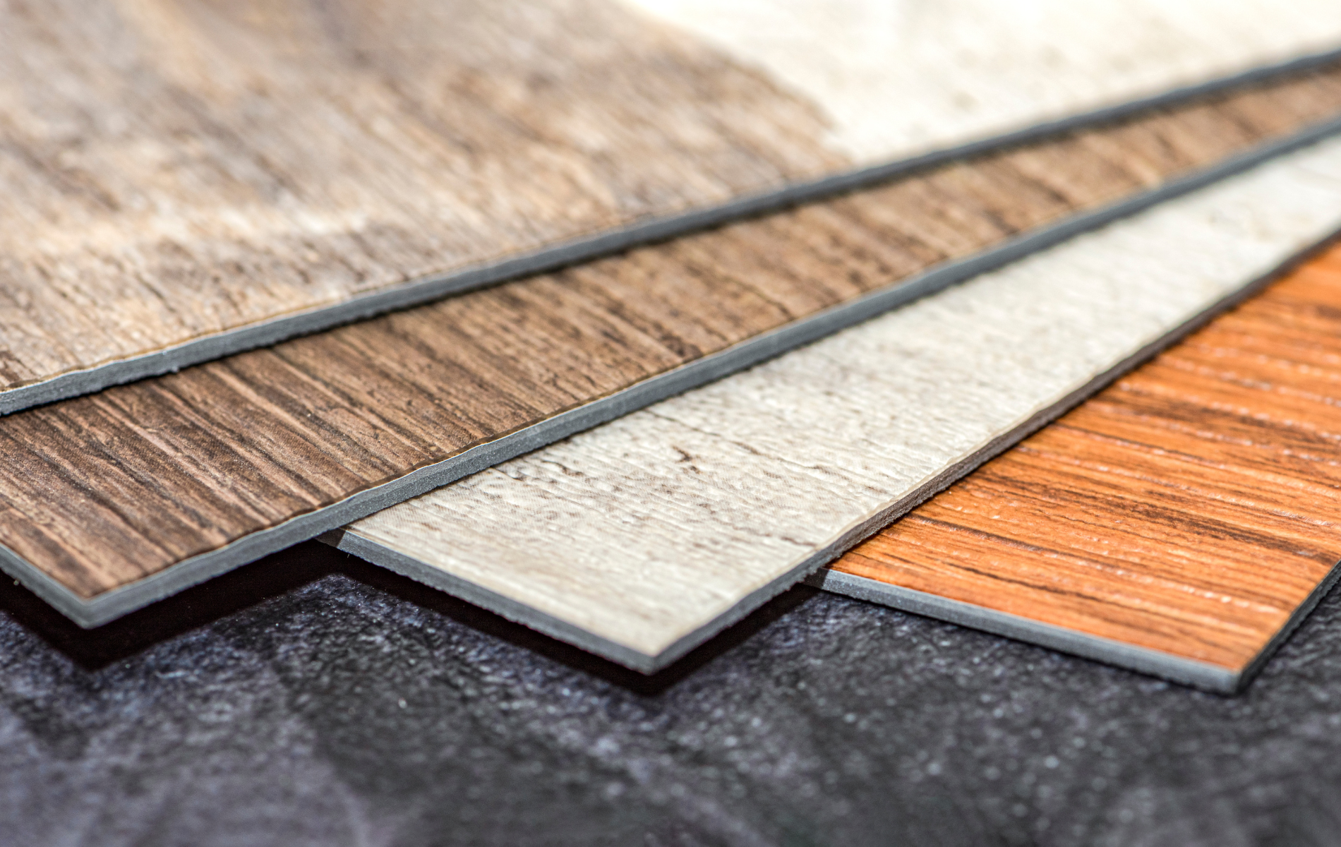 Four wood-look flooring samples in shades of brown, tan, and orange, displayed on a dark surface.