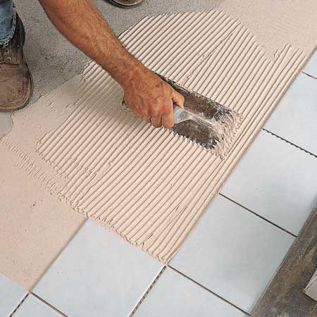 A man is spreading tile adhesive on a tiled floor with a spatula.