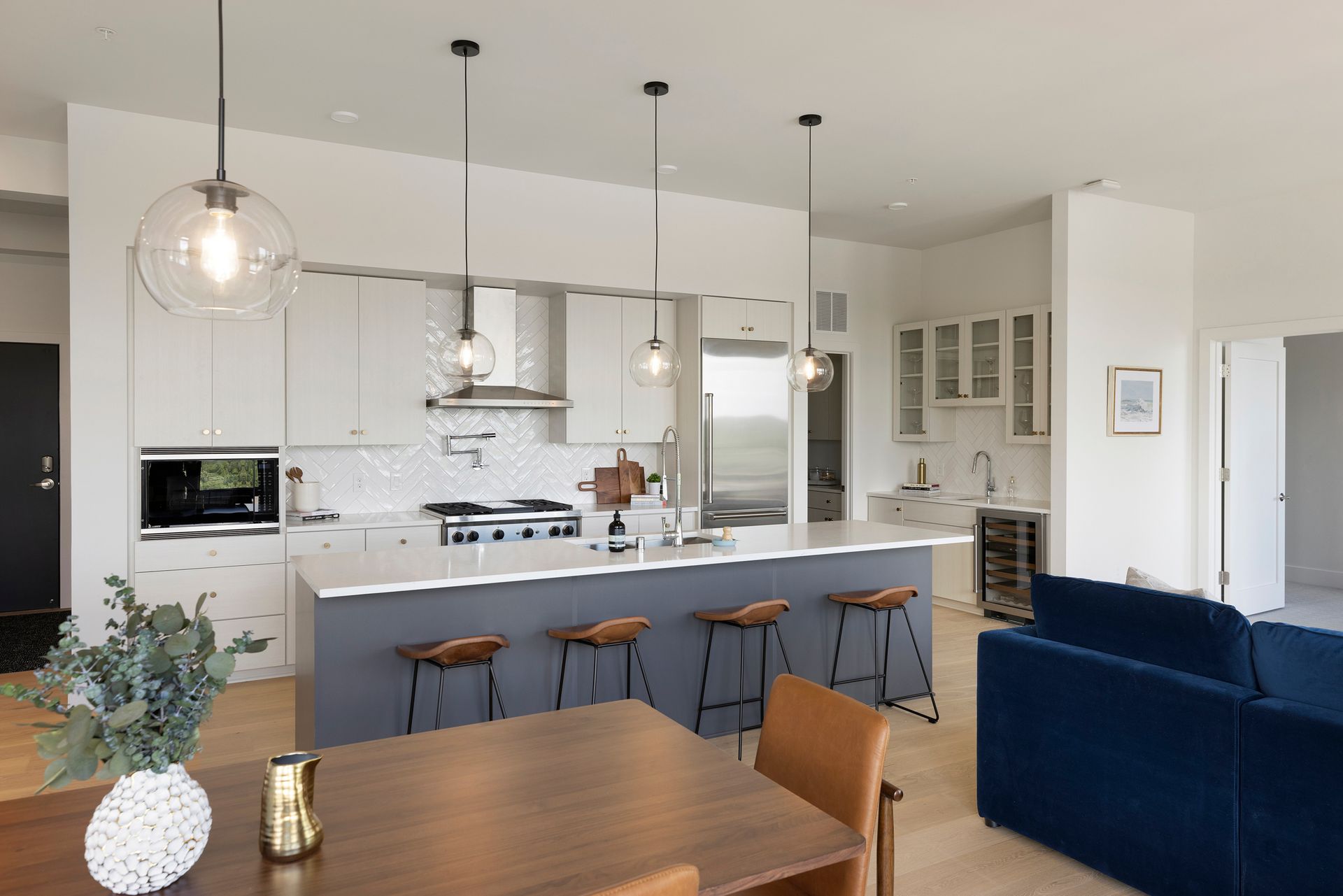 Modern kitchen with island and stools, open to dining area and living room; white cabinets, blue sofa.