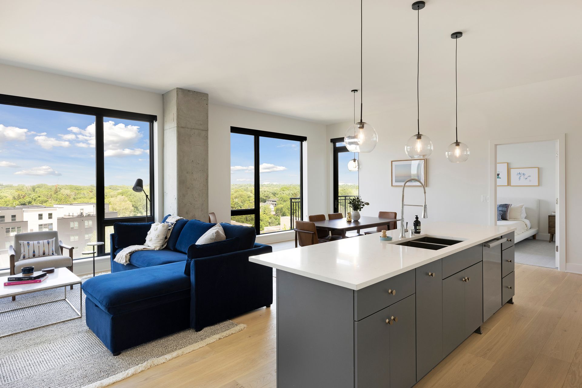 Modern open-plan living space with blue couch, gray kitchen island, and large windows overlooking trees.