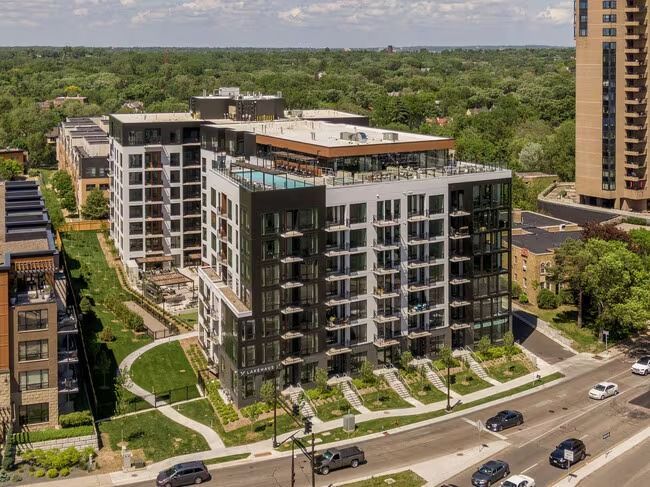 Modern apartment building with rooftop pool, surrounded by greenery and city street.