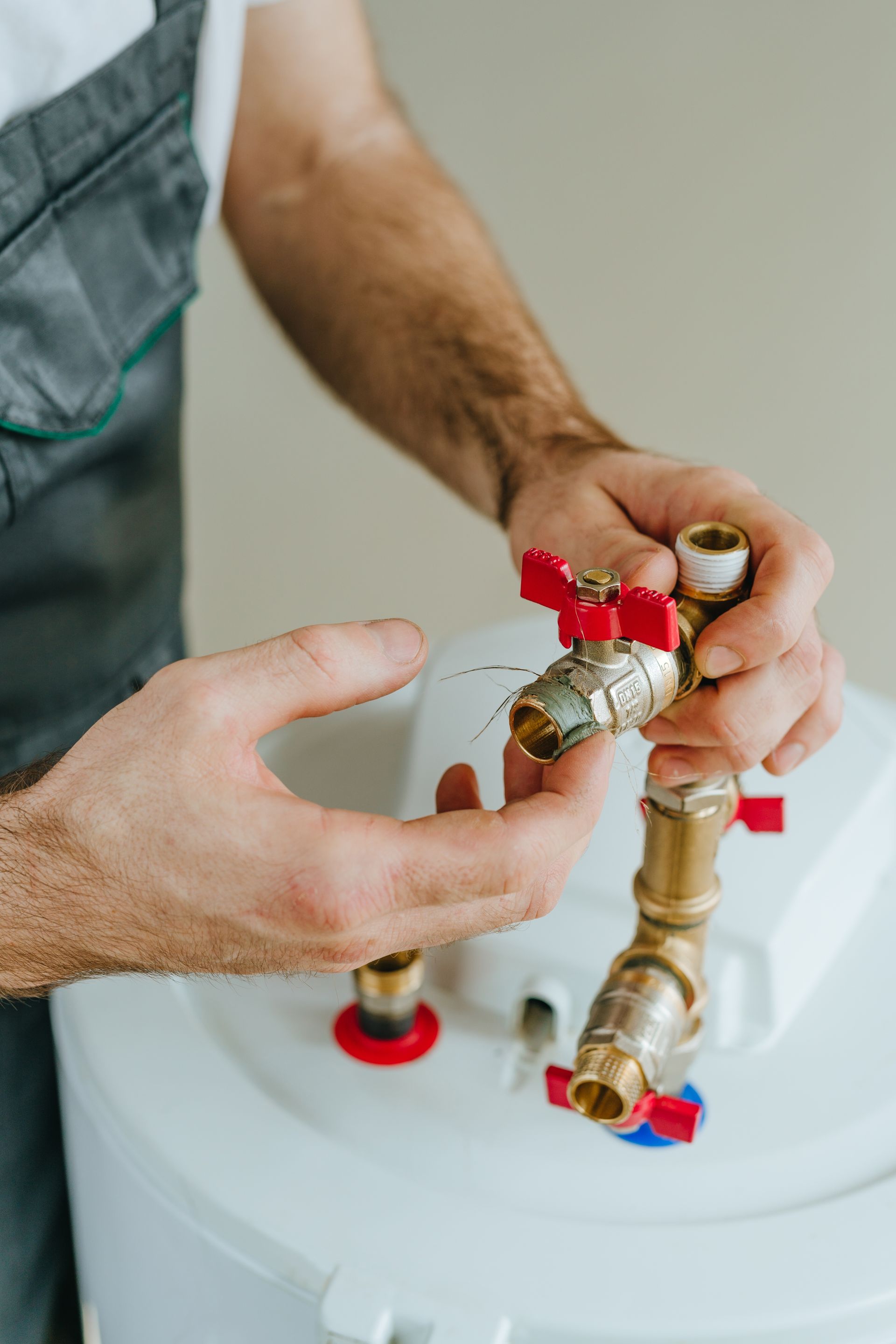 Plumber working on water valve, hands holding brass pipes with red and blue handles near a white tank.