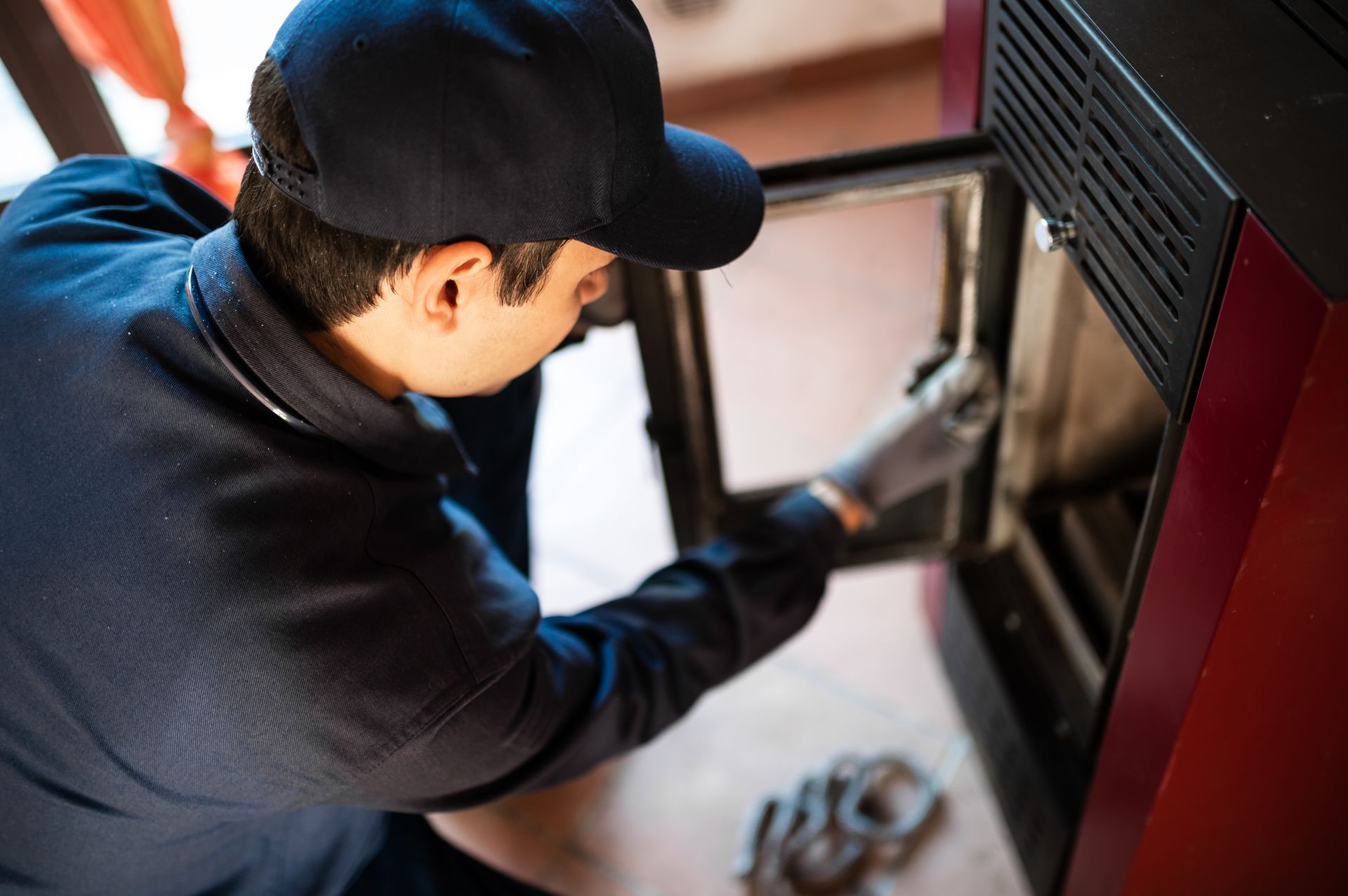 A person wearing a hat and gloves inspecting a red stove door.