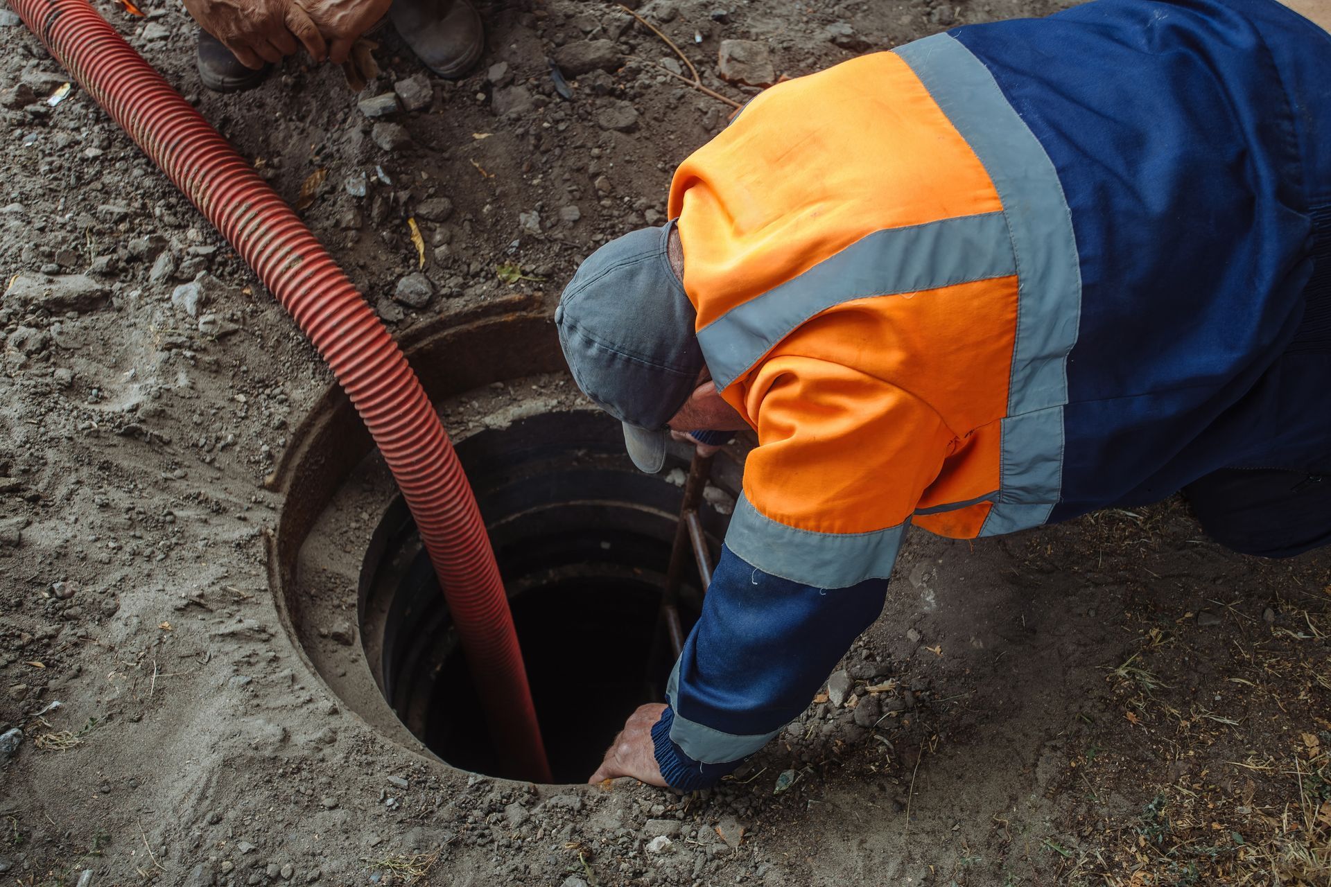 Man in safety vest leaning into open manhole, red hose nearby.
