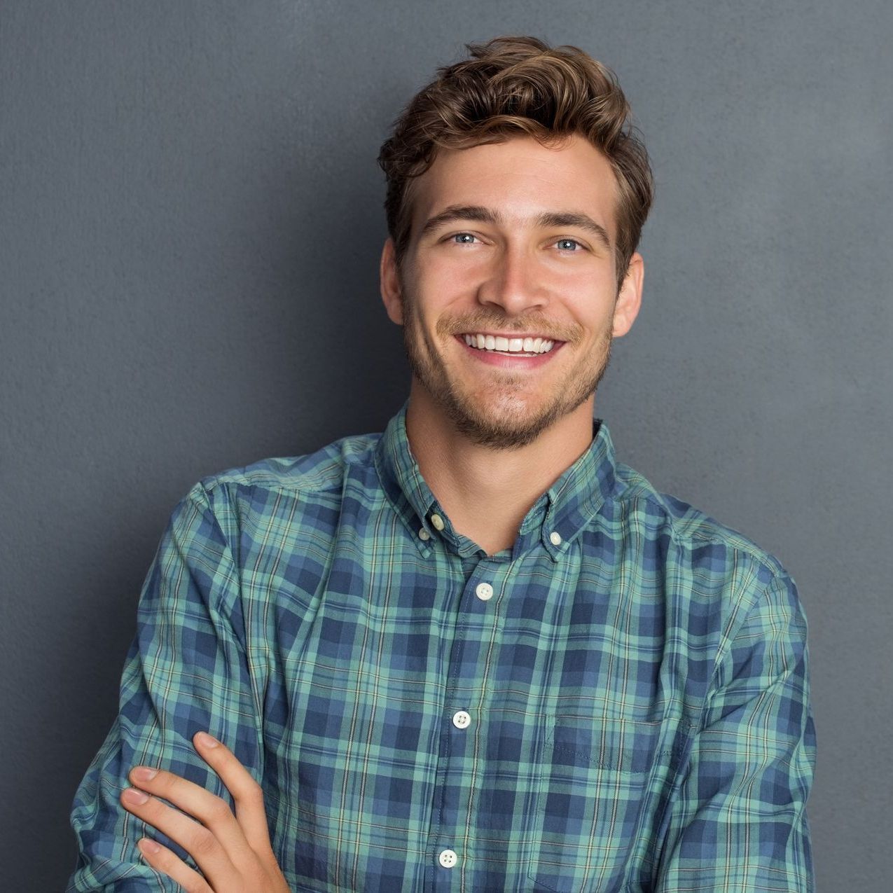 Smiling man with crossed arms wearing a blue and green plaid shirt against a gray background.