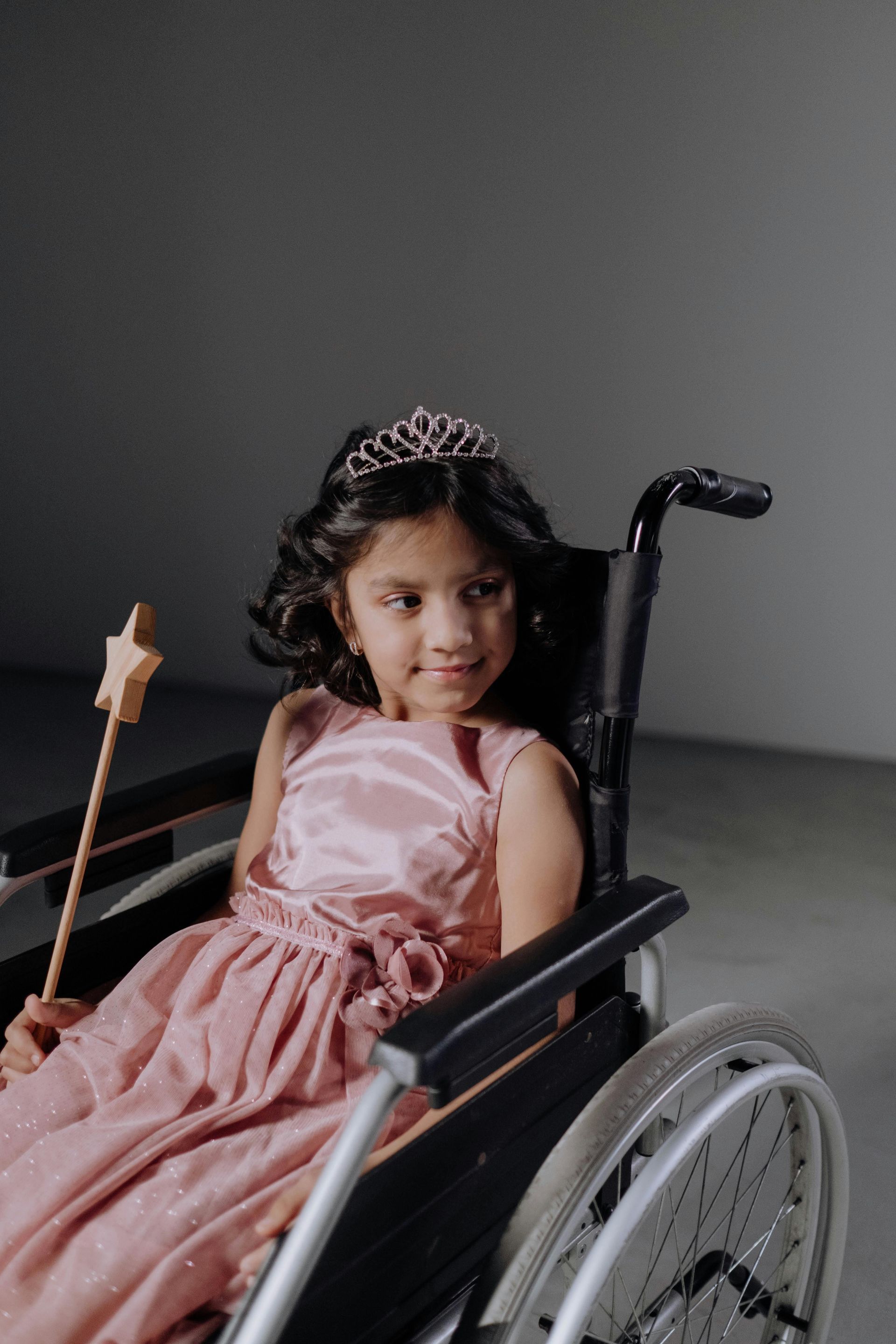 A child wearing a tiara and pink dress sits in a wheelchair, holding a star-topped wand.