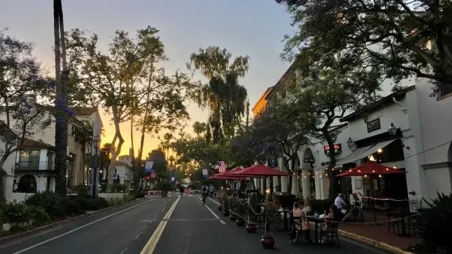 A street scene at sunset with outdoor restaurant seating under red umbrellas and trees along the sidewalk.